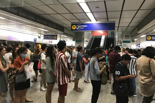 A busy underground MRT or BTS metro station in Bangkok, Thailand. Numerous passengers stand packed together on the platform, waiting for their train or moving toward the exits. Above the crowd hang blue signs reading „ทางออก" (Exit) in Thai script with English translation, along with arrows pointing toward the escalators. An escalator leads up to the exit. Fluorescent tubes on the ceiling brightly illuminate the area. On the right in the background, advertising panels for the Huawei Nova 4 smartphone can be seen. Many of those waiting are looking at their mobile phones. The people wear summer clothing — striped shirts, dresses, T-shirts, and sandals. One person on the left wears a light blue face mask. The floor is polished granite stone, and the station is modernly designed with metal cladding and a suspended ceiling.