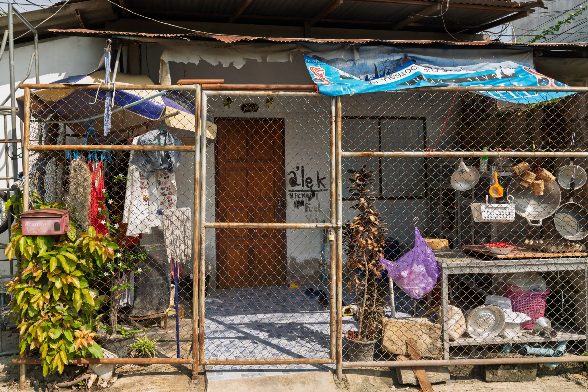 A modest residential dwelling in a Southeast Asian neighborhood, viewed from the front. The house has a brown wooden door with text scrawled on the wall beside it reading "alek" and other partially legible words. The entire front yard is enclosed by a chain-link fence with a bamboo and metal frame, secured with a padlock on the gate. Laundry and clothing items hang on the left side of the fence, alongside a red mailbox and lush green plants with white flowers. On the right side, various household items are displayed on shelves and hooks behind the fence, including metal pots, pans, a woven basket, a purple bag, and other kitchen utensils. A corrugated metal roof extends over the structure, supplemented by a blue tarpaulin banner stretched across the top for additional shade. Dried and potted plants sit inside the fenced area near the entrance. The overall scene reflects everyday life in a densely packed, working-class urban community.