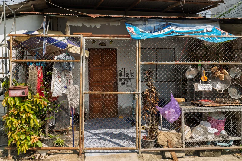 A modest residential dwelling in a Southeast Asian neighborhood, viewed from the front. The house has a brown wooden door with text scrawled on the wall beside it reading "alek" and other partially legible words. The entire front yard is enclosed by a chain-link fence with a bamboo and metal frame, secured with a padlock on the gate. Laundry and clothing items hang on the left side of the fence, alongside a red mailbox and lush green plants with white flowers. On the right side, various household items are displayed on shelves and hooks behind the fence, including metal pots, pans, a woven basket, a purple bag, and other kitchen utensils. A corrugated metal roof extends over the structure, supplemented by a blue tarpaulin banner stretched across the top for additional shade. Dried and potted plants sit inside the fenced area near the entrance. The overall scene reflects everyday life in a densely packed, working-class urban community.