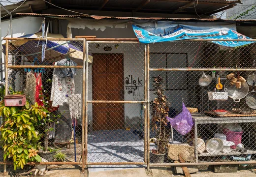 Frontansicht eines einfachen Wohnhauses in Südostasien, umgeben von einem Zaun aus Maschendraht mit einem Bambusrahmen und einer verschlossenen Gittertür in der Mitte. Hinter dem Zaun ist eine braune Holztür mit Graffiti-Schriftzügen zu sehen. Links hängen Kleidungsstücke und Wäsche an einer improvisierten Leine, darunter ein Kinderhemd und verschiedene Textilien. Rechts sind Küchenutensilien wie Töpfe, Pfannen, Siebe und Körbe an den Zaunstreben aufgehängt und auf Regalen abgestellt. Ein violetter Beutel hängt in der Mitte des Zauns. Über dem Eingangsbereich spannen sich zerschlissene Planen und Wellblechdächer als Wetterschutz. Links steht ein roter Briefkasten neben üppig grünen Pflanzen mit weißen Blüten. Auf dem Boden stehen Topfpflanzen, teilweise mit vertrockneten Blättern. Die Szene vermittelt ein Bild von bescheidenem, alltäglichem Leben in einer tropischen Umgebung mit viel improvisierter Nutzung des begrenzten Raums.