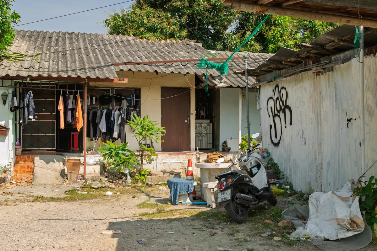 A simple, single-storey house in a Southeast Asian neighbourhood, likely in Thailand, with a corrugated tile roof and weathered concrete walls. Beneath an open canopy, numerous garments hang on hangers from a clothesline — orange, dark blue and grey shirts and jackets among them. In front of the house, a small tree with light green leaves stands next to a concrete step. An orange traffic cone and a low table covered with a cloth sit in the foreground. On the right, a black scooter with a Thai licence plate (336) is parked against a corrugated metal wall marked with black graffiti in Thai script. Large white bags of rubbish lie at the right edge of the frame. The scene shows typical features of a modest neighbourhood, with lush tropical vegetation in the background, green netting over the roof, and everyday items scattered around the outdoor area.