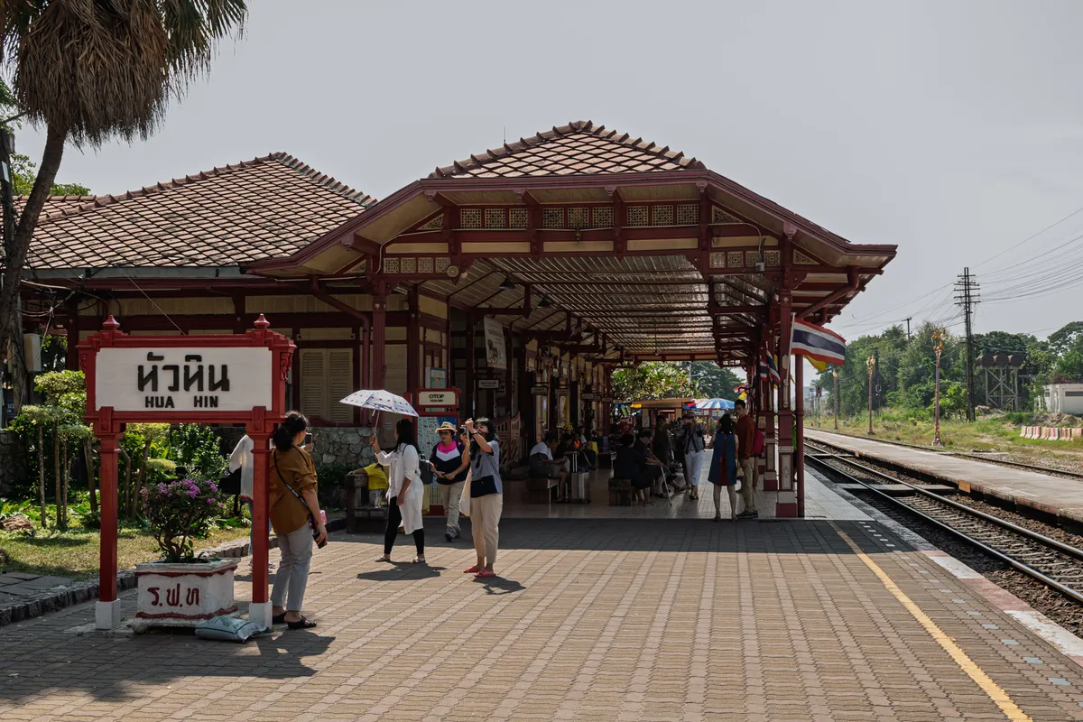 Hua Hin's historic railway station in Thailand with its distinctive red-and-cream wooden architecture in Victorian-Thai style. In the foreground stands a red-framed station sign reading "หัวหิน" (Hua Hin) in Thai and Latin script. Several tourists stand on the paved platform, some taking photos; one holds a parasol. The building has a tiered tiled roof with intricate wooden ornaments and open waiting areas beneath a long canopy. Tracks run along the platform on the right, and a Thai flag flies from a pillar. Trees, power poles and a signal bridge are visible in the background. To the left of the sign is a small, tidy garden with flowering shrubs and a palm.