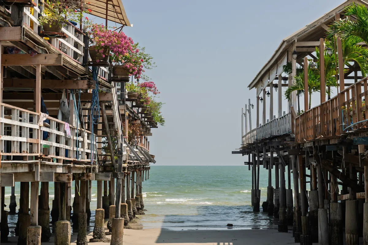 Two wooden stilt houses frame a narrow passage that opens onto the calm, turquoise sea and bright sky. The buildings rest on numerous wooden posts anchored in the sandy beach and shallow water. The structure on the left is multi-storey, with weathered white and brown wooden planks, hanging laundry, blue ropes and lush pink bougainvillea blooming in flower pots. The structure on the right has a gabled roof, railings and tropical palms. Between the buildings, gentle surf washes over the wet sand. The scene evokes a tropical coastal village, likely in Southeast Asia, in clear, slightly hazy daylight.