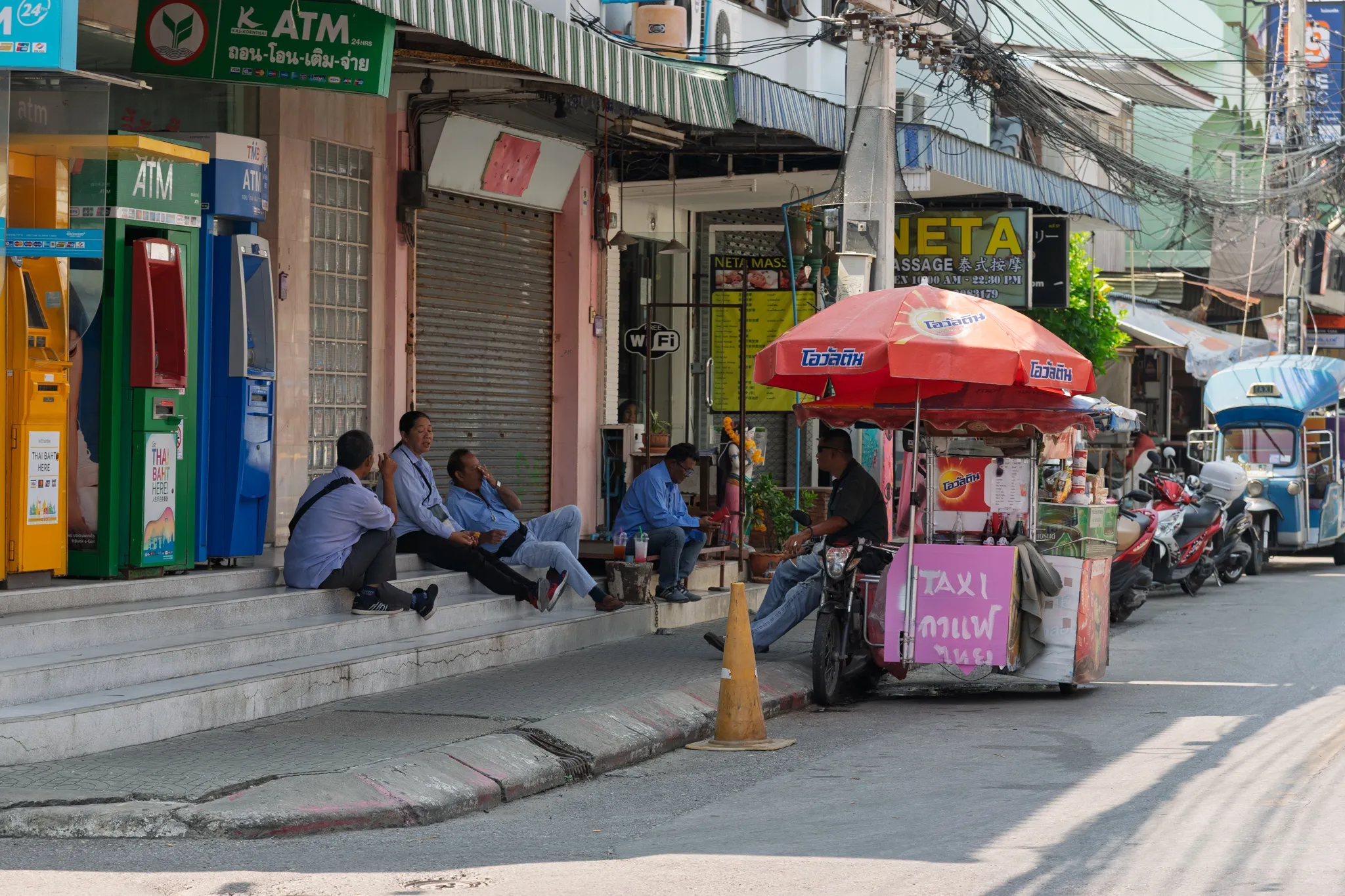 A bustling Thai street scene showing several men in blue uniforms, likely taxi or tuk-tuk drivers, sitting on concrete steps outside a row of shophouses, chatting and relaxing. To the right, a street vendor operates a mobile food and drink cart mounted on a motorcycle, shaded by a red Ovaltine-branded umbrella. The cart has a pink sign with Thai text reading "TAXI" and other Thai words. On the left side of the scene, colorful ATM machines from various Thai banks (Kasikorn, TMB, and others) are lined up along the building facade, with signs in Thai script. Behind the men, shuttered storefronts and a massage parlor called "NETA Massage" with Chinese characters are visible. The street is lined with parked motorcycles and a blue tuk-tuk can be seen further down the road. Overhead, a tangle of electrical wires stretches across utility poles, typical of Thai urban areas. A yellow traffic cone sits on the curb near the vendor cart.