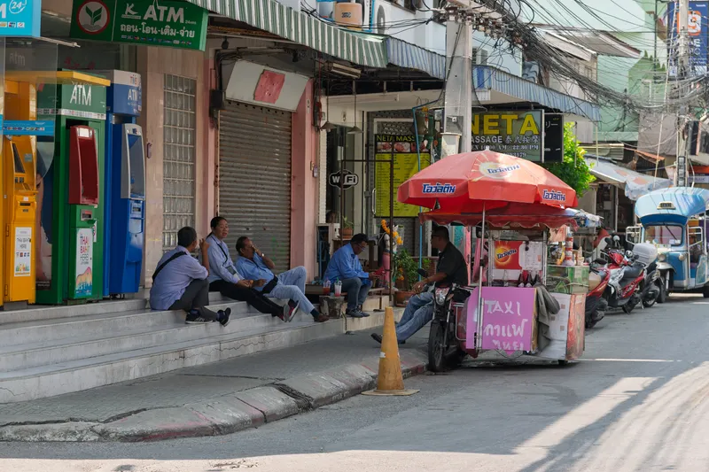 A bustling Thai street scene showing several men in blue uniforms, likely taxi or tuk-tuk drivers, sitting on concrete steps outside a row of shophouses, chatting and relaxing. To the right, a street vendor operates a mobile food and drink cart mounted on a motorcycle, shaded by a red Ovaltine-branded umbrella. The cart has a pink sign with Thai text reading "TAXI" and other Thai words. On the left side of the scene, colorful ATM machines from various Thai banks (Kasikorn, TMB, and others) are lined up along the building facade, with signs in Thai script. Behind the men, shuttered storefronts and a massage parlor called "NETA Massage" with Chinese characters are visible. The street is lined with parked motorcycles and a blue tuk-tuk can be seen further down the road. Overhead, a tangle of electrical wires stretches across utility poles, typical of Thai urban areas. A yellow traffic cone sits on the curb near the vendor cart.