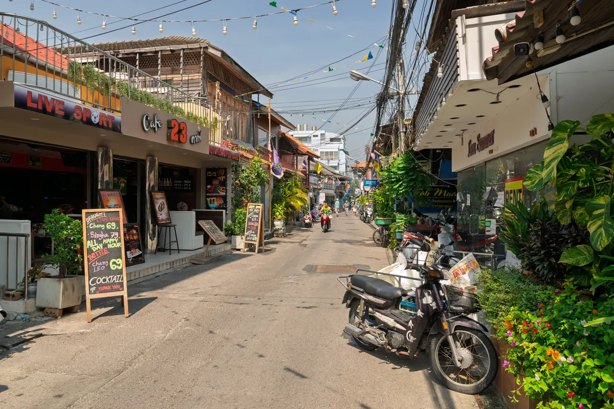 A narrow street in Thailand lined with bars, cafés and restaurants on both sides. On the left is an establishment marked "Live Sport" and "Cafe 23", with a chalkboard out front advertising Chang and Singha beer as well as cocktails. Motorbikes and scooters are parked along the right-hand kerb, surrounded by lush tropical plants and colourful flowers. String lights, colourful bunting and a tangle of power cables hang above the street. In the background, a person rides a red scooter while pedestrians stroll down the lane. The buildings are a mix of traditional Thai wooden architecture and modern façades. The sky is bright and slightly hazy, suggesting a warm, tropical time of day.