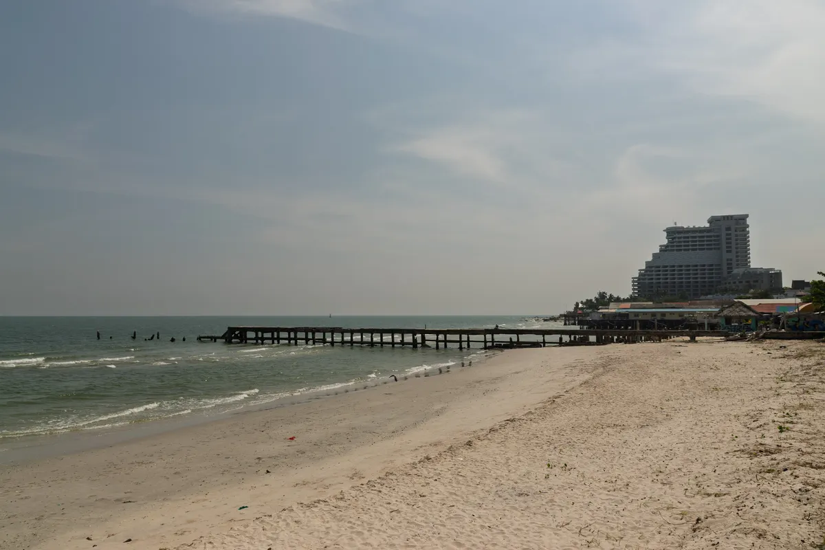 A broad, empty sandy beach with fine, pale sand stretches along a calm coastline. Gentle waves roll onto the shore. A long, dark wooden pier extends from the shore into the sea, with some of its pilings weathered and partly broken. On the right rises a striking multi-storey high-rise hotel of modern design, behind a row of smaller buildings — thatched beach huts and low commercial structures. Palm trees are visible between the buildings. The sky is lightly overcast and hazy, giving the whole image a muted, calm atmosphere. The sea stretches to the horizon in a greyish-green hue.
