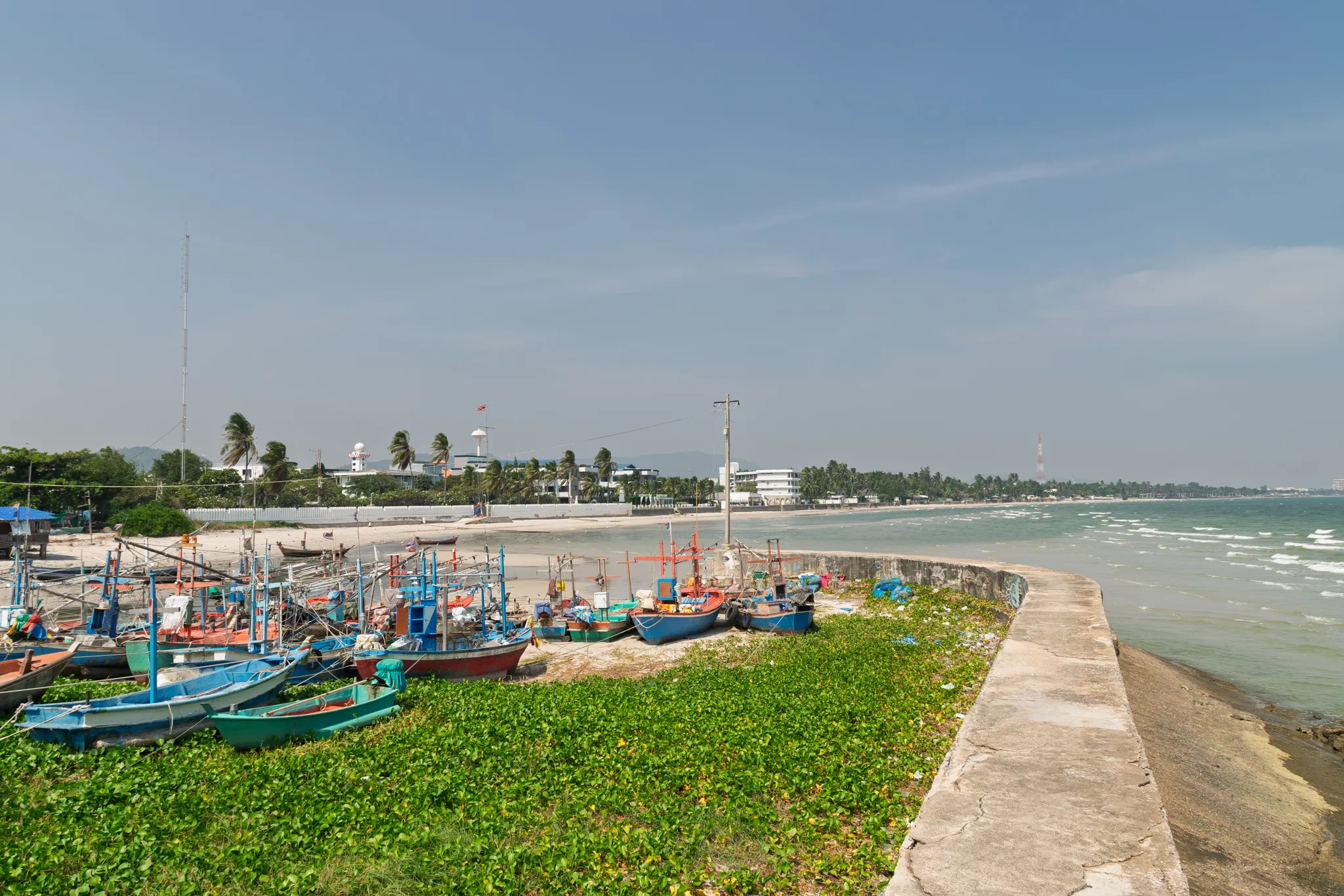 A coastal fishing village scene with numerous colorful wooden fishing boats — painted in shades of blue, green, red, and teal — beached on a sandy shore covered with lush green beach morning glory vines. A curved concrete seawall runs along the right side, separating the shore from the turquoise ocean with gentle white-capped waves. In the background, palm trees and coconut palms line the coastline, with low-rise white buildings, a water tower, a telecommunications tower on the left, and a tall red-and-white smokestack or tower visible in the distance on the right. Distant mountains are faintly visible under a hazy blue sky with thin wispy clouds. The boats are equipped with fishing gear, poles, and nets, suggesting an active local fishing community, likely in Southeast Asia, possibly Thailand.