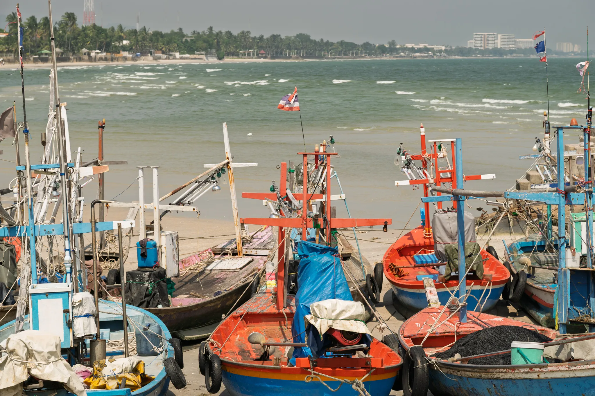 Colorful Thai fishing boats moored on a sandy beach along the coastline. The boats, painted in vibrant shades of red, blue, orange, and white, are equipped with wooden cross-shaped mast structures, ropes, tarps, fishing gear, and small flags, including a Thai national flag fluttering from one of the masts. The vessels sit on the sand near the waterline, with choppy green-gray ocean waves breaking in the background. Along the distant shoreline, palm trees, low buildings, and a large multi-story hotel or resort building are visible under an overcast hazy sky. The scene captures a traditional Thai fishing village atmosphere with well-used, weathered boats clustered together.