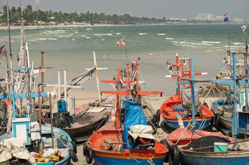 Colorful Thai fishing boats moored on a sandy beach along the coastline. The boats, painted in vibrant shades of red, blue, orange, and white, are equipped with wooden cross-shaped mast structures, ropes, tarps, fishing gear, and small flags, including a Thai national flag fluttering from one of the masts. The vessels sit on the sand near the waterline, with choppy green-gray ocean waves breaking in the background. Along the distant shoreline, palm trees, low buildings, and a large multi-story hotel or resort building are visible under an overcast hazy sky. The scene captures a traditional Thai fishing village atmosphere with well-used, weathered boats clustered together.