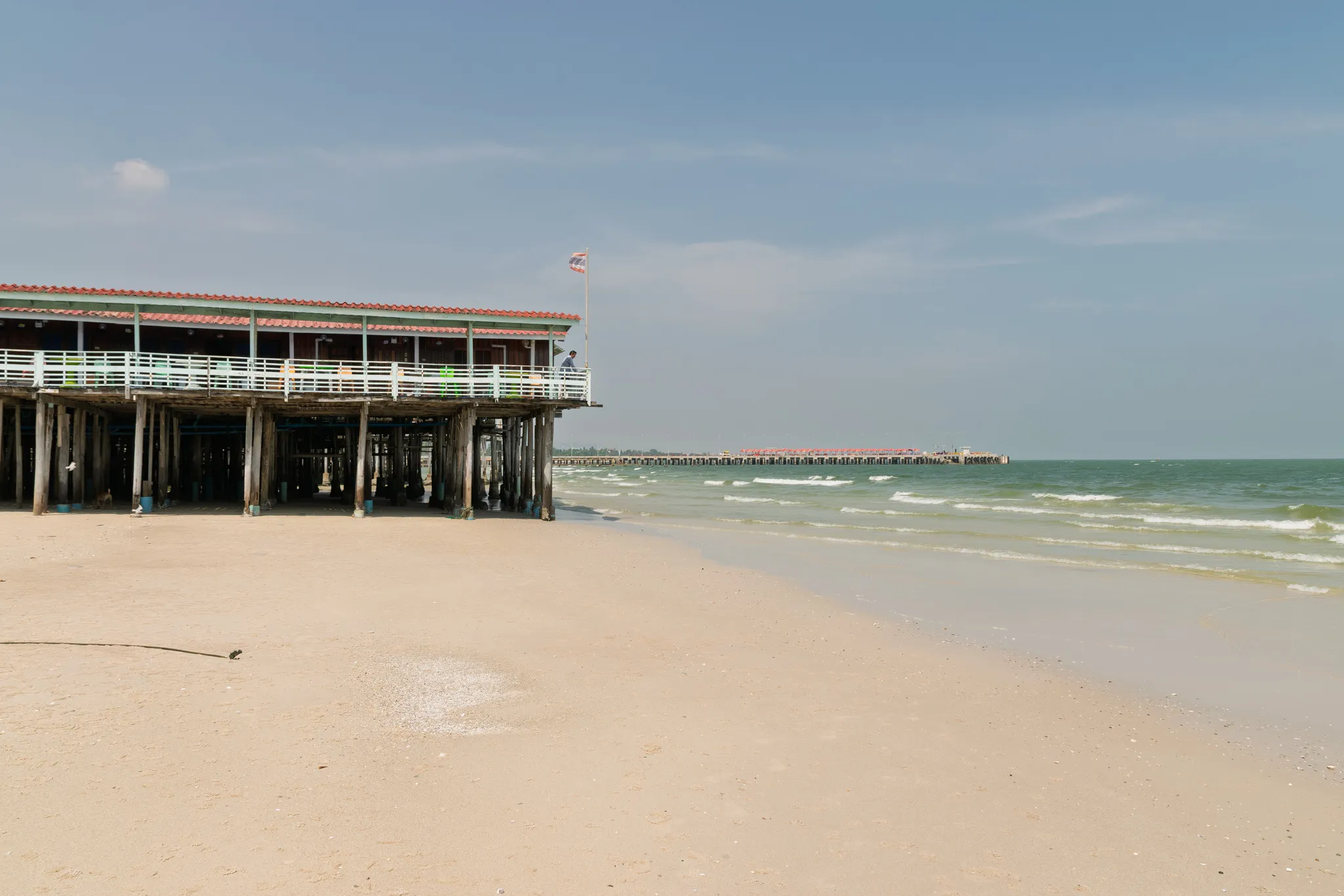 A wooden pier with a red-tiled roof structure stands on weathered timber pilings along a sandy beach. The building features white railings and a green-trimmed facade, with a flag flying from a pole on top. Gentle waves lap at the shoreline where the wet sand meets the calm, greenish ocean water. In the background, another pier or jetty extends into the sea. The sky is mostly clear with light wispy clouds, and the beach appears quiet and largely empty. Shells and small debris are scattered across the sand near the waterline.