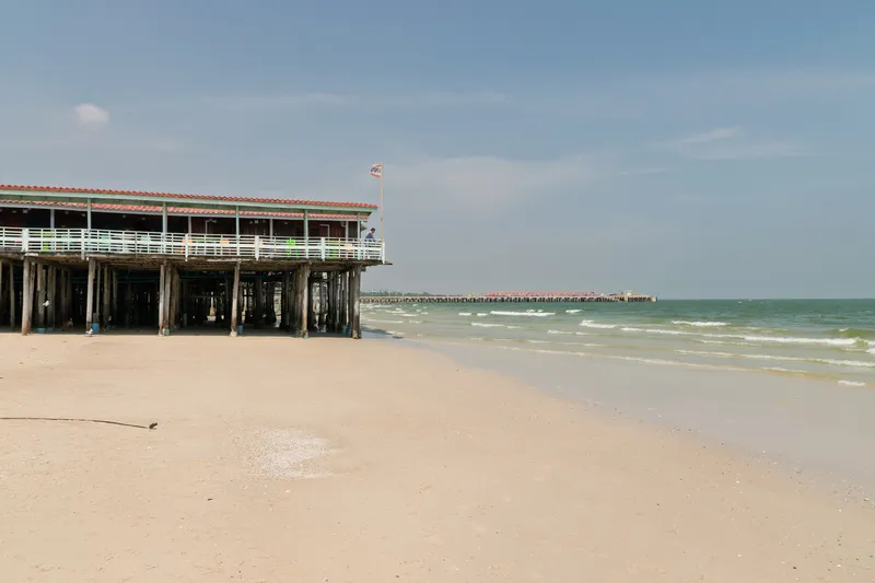 A wooden pier with a red-tiled roof structure stands on weathered timber pilings along a sandy beach. The building features white railings and a green-trimmed facade, with a flag flying from a pole on top. Gentle waves lap at the shoreline where the wet sand meets the calm, greenish ocean water. In the background, another pier or jetty extends into the sea. The sky is mostly clear with light wispy clouds, and the beach appears quiet and largely empty. Shells and small debris are scattered across the sand near the waterline.