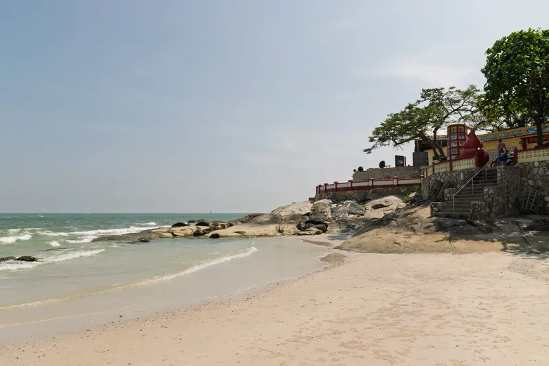 A sandy beach along the coast with gentle turquoise waves lapping at the shore. On the right side, large natural rock formations extend from the beach into the water, leading up to a colorful Vietnamese or Southeast Asian temple or shrine built on elevated ground. The temple features a yellow wall with decorative tile work, red ornamental elements including large gourd-shaped structures, a stone staircase with metal railings, and a red-and-white balustrade. Mature tropical trees with broad green canopies shade the temple area. A person on a motorbike is visible near the temple entrance. The sky is hazy and pale blue, and the sea stretches to the horizon on the left. The beach is mostly empty with fine light-colored sand scattered with small shells and pebbles.