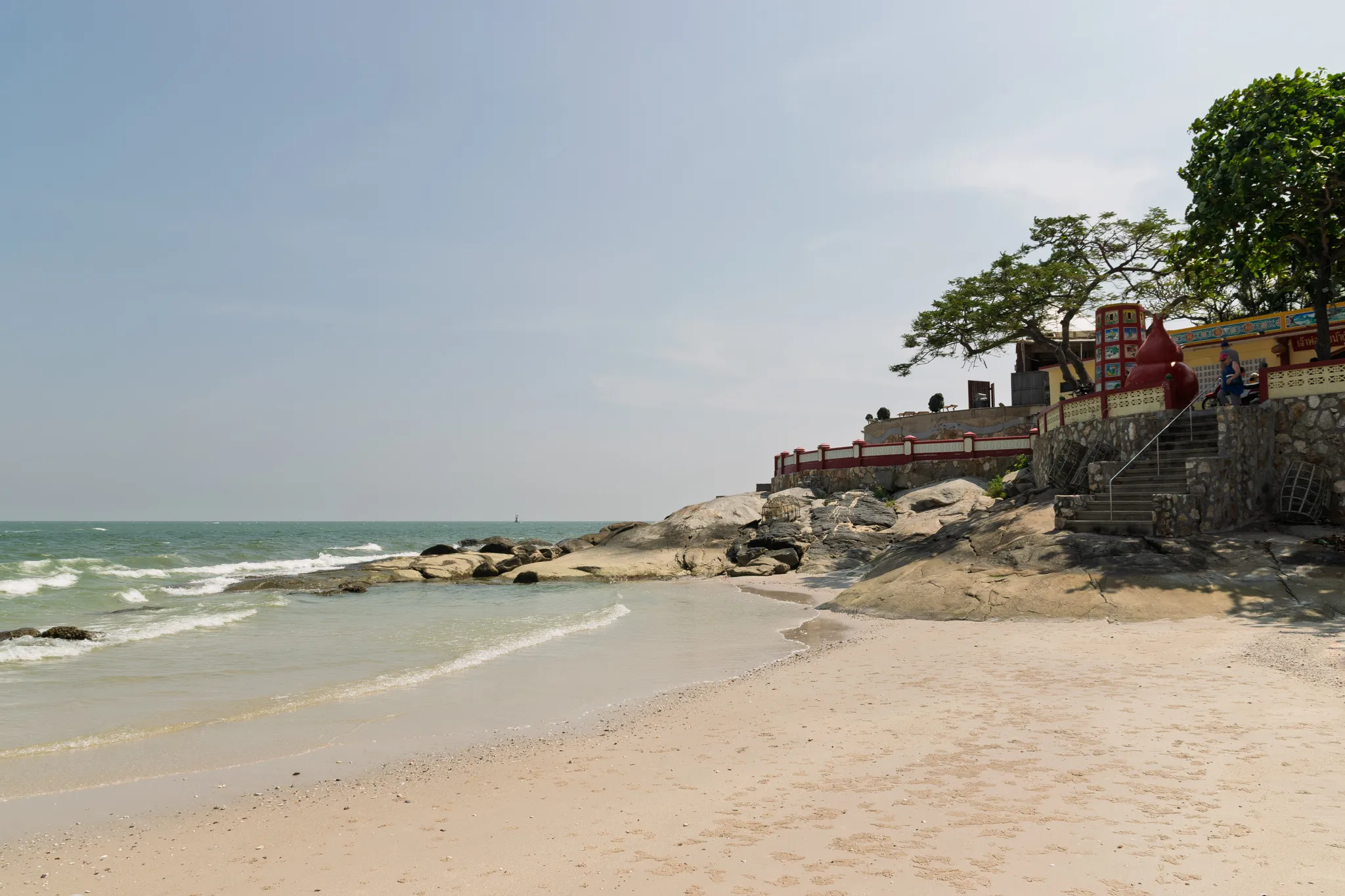 A sandy beach along the coast with gentle turquoise waves lapping at the shore. On the right side, large natural rock formations extend from the beach into the water, leading up to a colorful Vietnamese or Southeast Asian temple or shrine built on elevated ground. The temple features a yellow wall with decorative tile work, red ornamental elements including large gourd-shaped structures, a stone staircase with metal railings, and a red-and-white balustrade. Mature tropical trees with broad green canopies shade the temple area. A person on a motorbike is visible near the temple entrance. The sky is hazy and pale blue, and the sea stretches to the horizon on the left. The beach is mostly empty with fine light-colored sand scattered with small shells and pebbles.