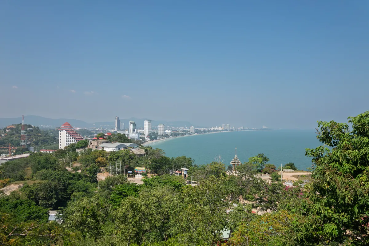 A panoramic view from an elevated vantage point over a tropical coastal town, likely Hua Hin in Thailand. In the foreground, lush green vegetation with dense tree canopies stretches out. On a hill to the left, buildings with striking red Asian-style roofs are visible, including a temple-like structure. In the middle, a multi-storey pagoda rises among the trees. Beyond that, the city skyline extends with several high-rises and hotels along a curving bay with a long sandy beach. The calm, turquoise sea reaches to the horizon, where it merges into the slightly hazy, light blue sky. Gentle mountain ranges are faintly visible in the background. A transmission tower and a few construction cranes hint at the region's urban development.