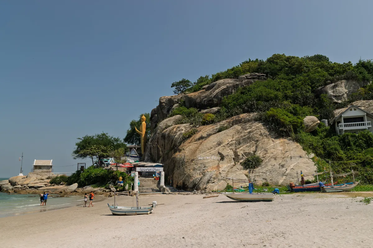 A tropical sandy beach on a rocky coast in Thailand, dominated by a large granite boulder overgrown with green vegetation. At its base stands a Buddhist temple with a golden standing Buddha statue and an entrance with stairs, marked in Thai script. On a rock ledge to the left stands a small white building with a pointed roof. Several fishing boats and small wooden boats lie on the pale sand. A few visitors, adults and children, stroll at the water's edge. On the right, a multi-storey residential building is visible on the slope. The sky is clear and blue, the sea calm and turquoise.