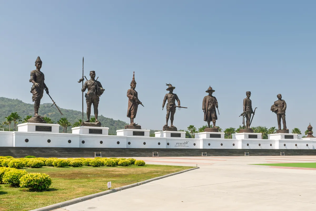 Seven large bronze statues of Thai kings stand in a row on white stone pedestals against a clear, slightly hazy sky. The figures are depicted in traditional Thai dress and armour, some equipped with spears, swords and other weapons. Each statue stands on its own plinth with a dark information plaque. In the background, a green, plant-covered hill rises with palm trees, while the foreground shows a well-kept lawn with low-cut yellow-green hedges and a broad paved path. The site is part of Rajabhakti Park in Hua Hin, Thailand, dedicated to the seven great kings of Thai history. A sign in Thai script is mounted on the white wall below the middle statue.