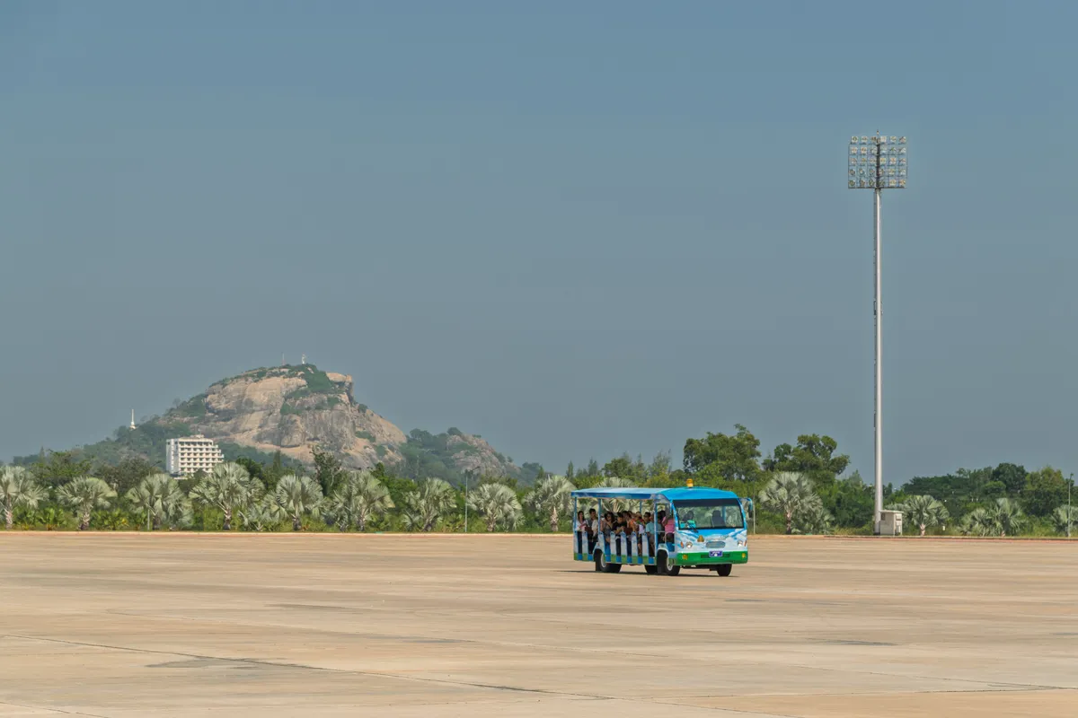 A blue-green-and-white open-sided tourist bus full of passengers drives across a broad, empty concrete plaza. In the background, a striking rocky peak with sparse vegetation rises, with a multi-storey building at its base. The landscape is lined with tropical palms and trees. On the right, a tall floodlight mast stretches upward. The sky is hazy and slightly grey-blue, suggesting tropical, humid weather. The scene feels like a large plaza or site in Southeast Asia, possibly Myanmar (Naypyidaw), given the characteristic vastness of the space and the vegetation.