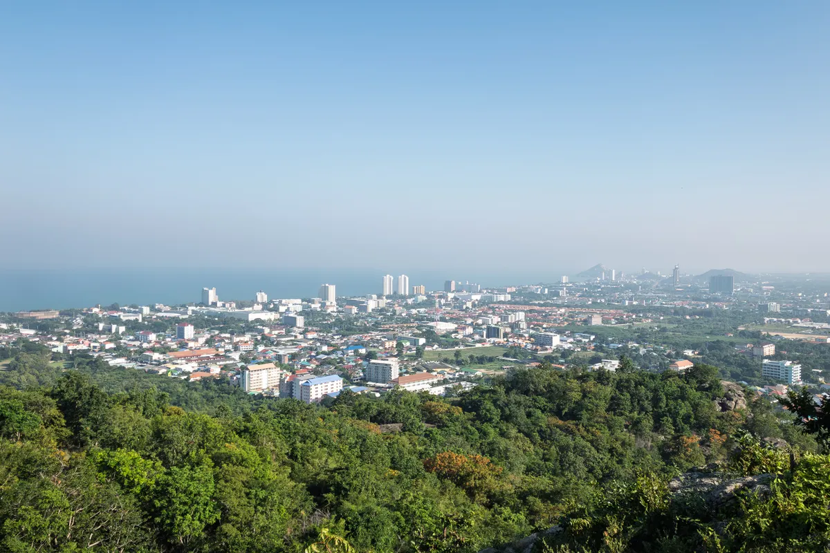 A panoramic view from a wooded hill over a sprawling coastal town in a tropical setting. In the foreground stretches a dense, deep green deciduous forest with scattered rock formations. Beyond lies a broad cityscape with countless low residential buildings, terraced-house estates with red and orange roofs, and several white high-rises and skyscrapers. On the horizon, the calm sea shimmers under a slightly hazy, light blue sky. On the right, mountainous peaks rise from the haze. The scene is reminiscent of a Southeast Asian coastal town, possibly Hua Hin in Thailand, captured in clear weather with soft morning light.