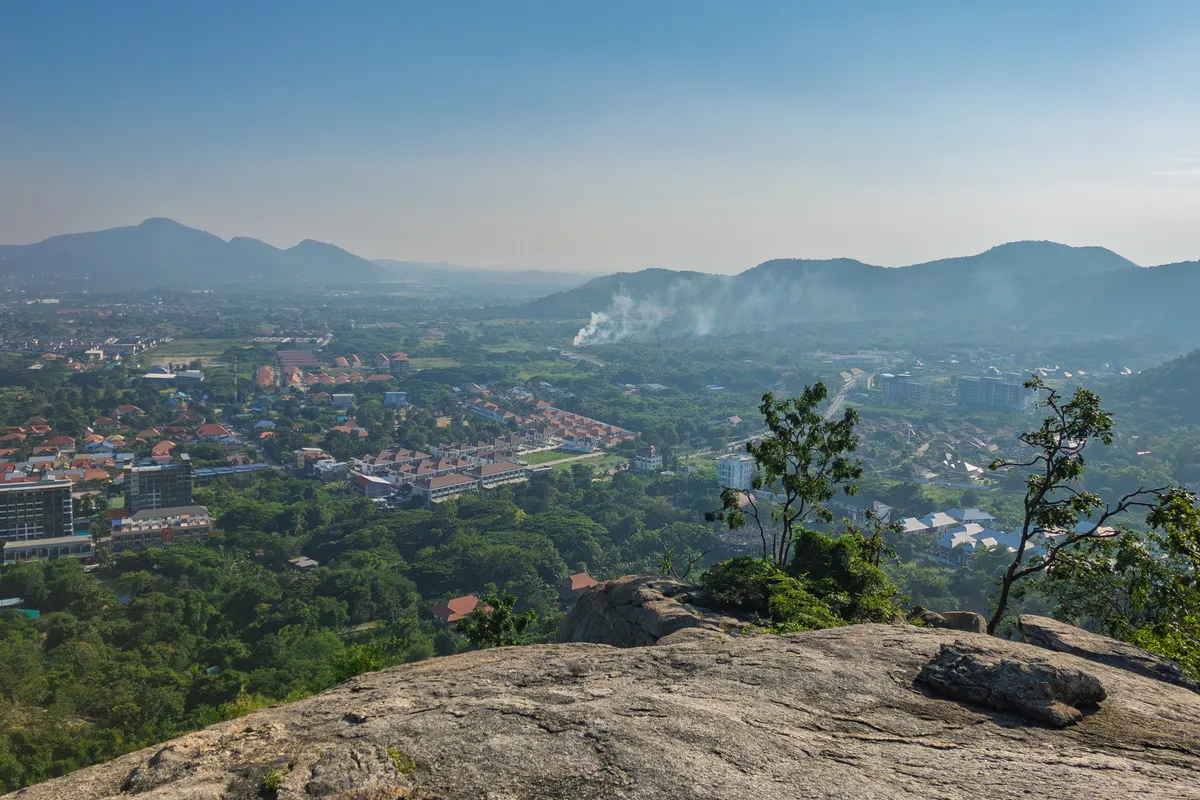 A panoramic view from a rocky lookout over a sprawling urban and natural landscape in Southeast Asia. In the foreground, a large flat granite slab stretches out, with a small tree growing at its edge. Below spreads a green, densely vegetated plain dotted with countless residential estates with orange and red tiled roofs, modern buildings and the occasional high-rise. In the middle distance, light smoke rises, possibly from slash-and-burn or a fire. In the background, several mountain ranges extend in various shades of blue, fading progressively into the haze of the humid tropical atmosphere. The sky is slightly hazy with a soft blue, typical of tropical regions in the morning hours. The lush vegetation and architecture suggest a location in Thailand, possibly the area around Hua Hin or Pattaya.