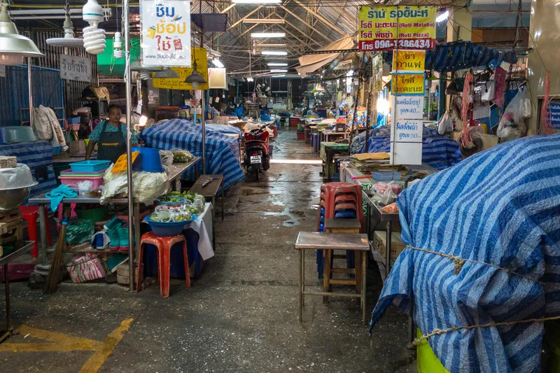 A Thai street market in the evening or early morning, mostly closed down with stalls covered by blue and white striped tarpaulins. A narrow wet concrete aisle runs through the center of the covered market, with a red motorcycle parked in the middle distance. On the left side, a woman vendor in an apron stands behind her stall, which displays bowls of fresh vegetables and ingredients on tables with colorful plastic stools and baskets underneath. Signs in Thai script hang from poles and above stalls, including advertisements for "Roti Mataba" and "Roti Pizza" with a phone number (096-1386434), along with menu prices of 40 and 50 baht. On the right side, wooden stools and small tables sit in front of covered food stalls, with yellow and blue price signs advertising tea and coffee. The market has a corrugated metal roof with fluorescent and CFL lights illuminating the space. The damp floor and quiet atmosphere suggest the market is either closing for the night or has not yet fully opened.