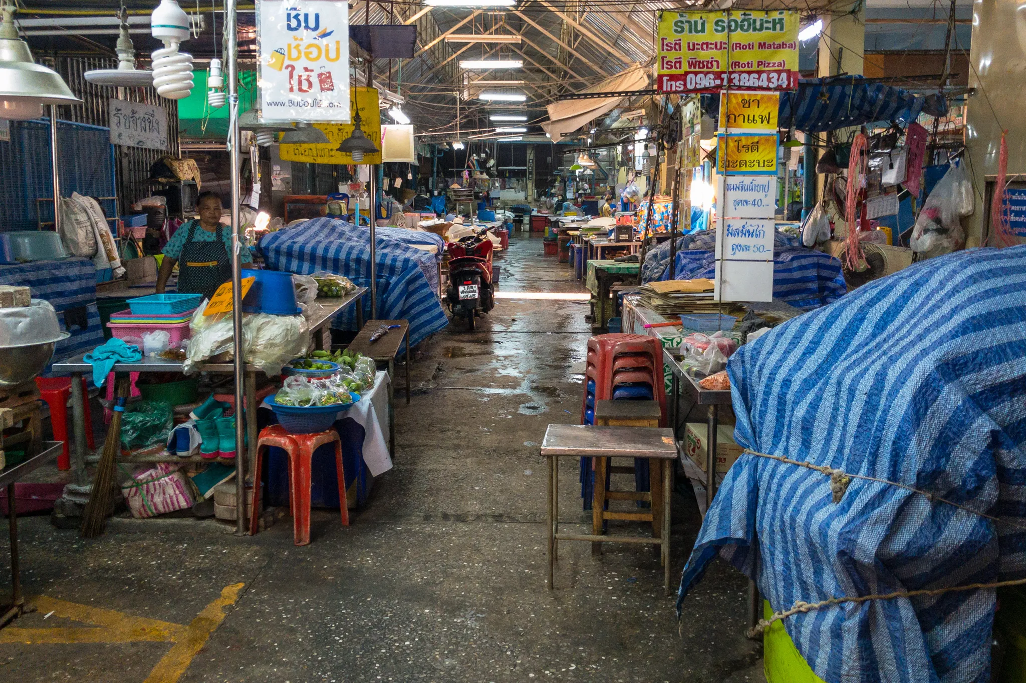 A Thai street market in the evening or early morning, mostly closed down with stalls covered by blue and white striped tarpaulins. A narrow wet concrete aisle runs through the center of the covered market, with a red motorcycle parked in the middle distance. On the left side, a woman vendor in an apron stands behind her stall, which displays bowls of fresh vegetables and ingredients on tables with colorful plastic stools and baskets underneath. Signs in Thai script hang from poles and above stalls, including advertisements for "Roti Mataba" and "Roti Pizza" with a phone number (096-1386434), along with menu prices of 40 and 50 baht. On the right side, wooden stools and small tables sit in front of covered food stalls, with yellow and blue price signs advertising tea and coffee. The market has a corrugated metal roof with fluorescent and CFL lights illuminating the space. The damp floor and quiet atmosphere suggest the market is either closing for the night or has not yet fully opened.