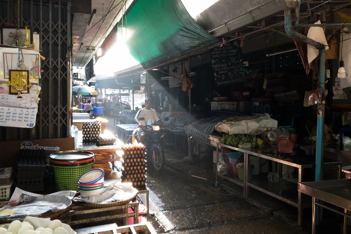 A helmeted motorcyclist rides through a narrow, shaded alley in a Thai market. Sunbeams fall dramatically from above through a gap between green tarps and canopies, lighting the wet floor and stacked egg cartons on the left. Colourful plastic baskets and bowls sit on a wooden table on the left, behind which a shop with a closed metal grille displays a framed picture of a Buddhist temple. On the right, stalls stretch out with stainless-steel tables, green bananas, plastic baskets and a chalkboard in Thai script. Water pipes and cables run along the ceiling. The atmosphere is damp and lively, typical of a traditional Thai fresh market in the early morning.