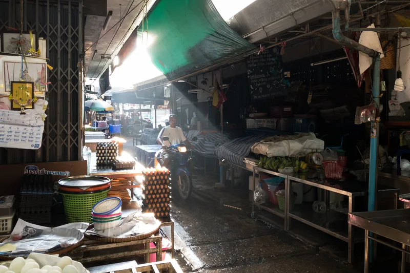 A narrow alleyway in a Thai wet market, bathed in dramatic rays of morning sunlight streaming through a green tarpaulin canopy overhead. A man on a motorcycle with its headlight on rides through the narrow passage between market stalls. On the left side, stacks of egg cartons filled with eggs sit on a surface, alongside colorful plastic baskets and woven trays. Newspapers are spread beneath the goods. A metal security gate with a diamond pattern is partially visible on the far left, with a framed picture of a Thai temple and a spirit house shrine hanging above. On the right side, a food stall area features metal shelving with green bananas, plastic baskets, and covered surfaces draped in striped cloth. A chalkboard menu with Thai script hangs on the wall, and a small Thai flag is visible. The wet ground reflects the light, and the overall atmosphere is moody and atmospheric with visible light beams cutting through the humid air.