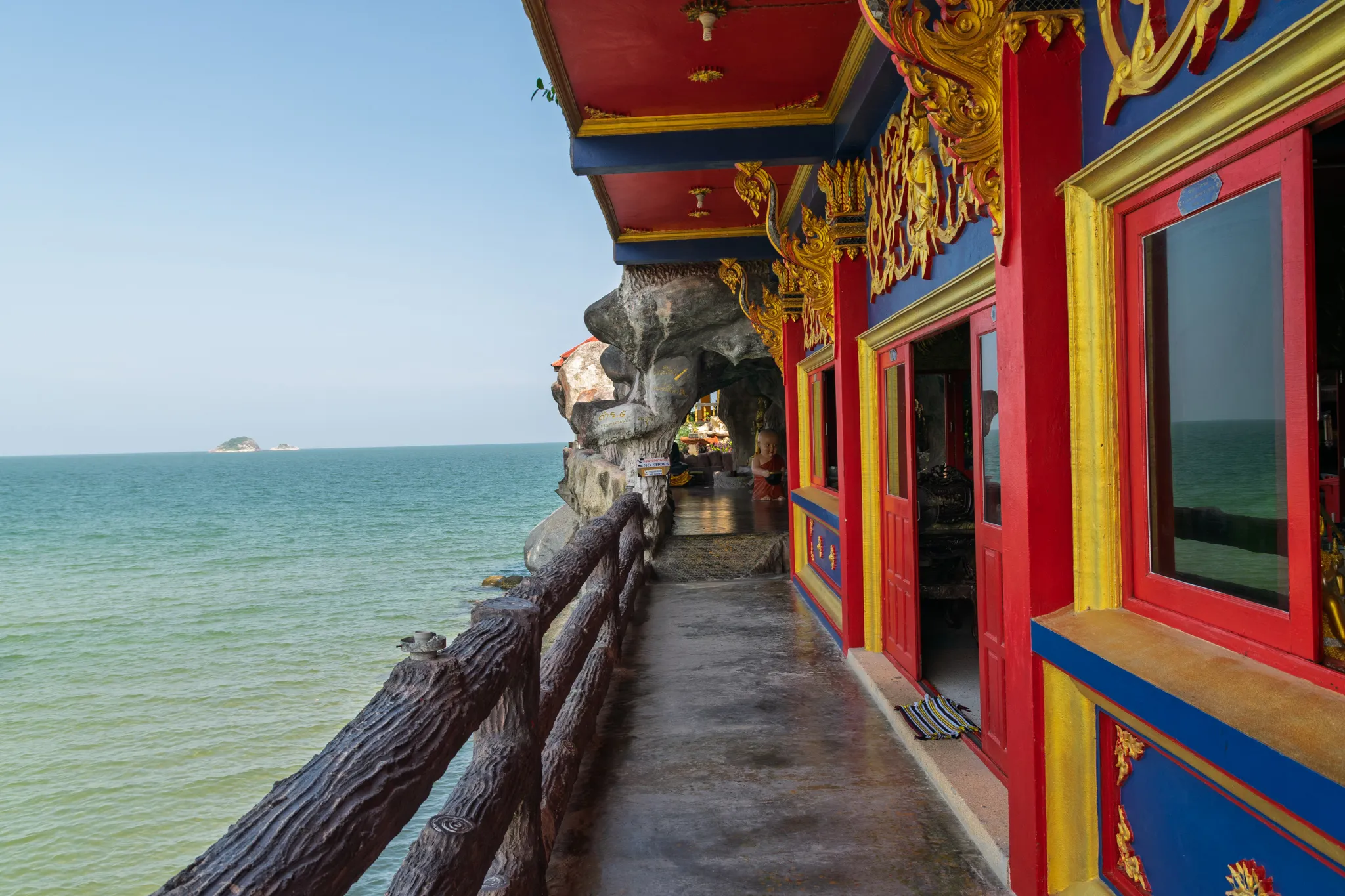 A colorful Buddhist temple built into a rocky cliff overlooking a calm, greenish sea. The temple features vibrant red, blue, and yellow painted walls with ornate golden gilded carvings and decorative motifs along the eaves and window frames. A narrow concrete walkway runs along the seaside edge of the temple, bordered by a rustic wooden railing made to resemble natural tree branches. The walkway leads toward a natural rock formation or cave area where Buddhist statues and offerings are visible in the background. Large glass windows and red doors line the temple wall. The sea stretches to the horizon under a hazy blue sky, with small islands visible in the distance. Sandals are placed neatly outside one of the temple doors.