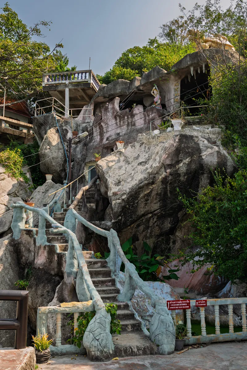 A steep stone staircase with ornamental tree-shaped concrete railings painted in pale blue-green leads up a large rocky hillside to a Thai Buddhist temple built into the natural rock formation. At the top of the stairs, a cave-like structure carved into the rock features a seated Buddhist figure, with Thai script visible above. The rock face is massive and dark gray, with lush tropical vegetation and trees surrounding the site. To the right, red signs with white text in both Thai and English read "Guan Im Buddha Posture 1000 Hands" and "Laansai." A white balustrade runs along the base of the stairway, and potted plants sit near the bottom. A building with a concrete balcony railing is partially visible at the upper left, built into the rock. The sky above is pale blue and slightly hazy, suggesting warm tropical weather.