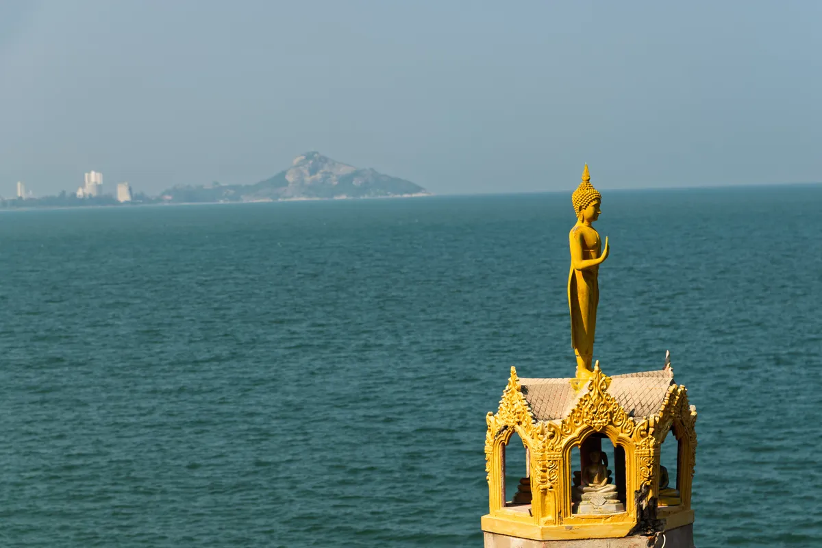 A golden, ornately decorated Buddhist temple shrine with a standing Buddha statue on the roof, one hand raised in a gesture of blessing. The shrine sits in the lower right of the frame and is richly adorned with delicate ornaments and typically Thai gable decorations. Inside the small pavilion sits another, smaller Buddha figure. In the background, a calm turquoise-green sea stretches to the horizon, where a mountainous coastline with a few high-rises is visible in the distance through the light haze. The sky is pale and slightly hazy.