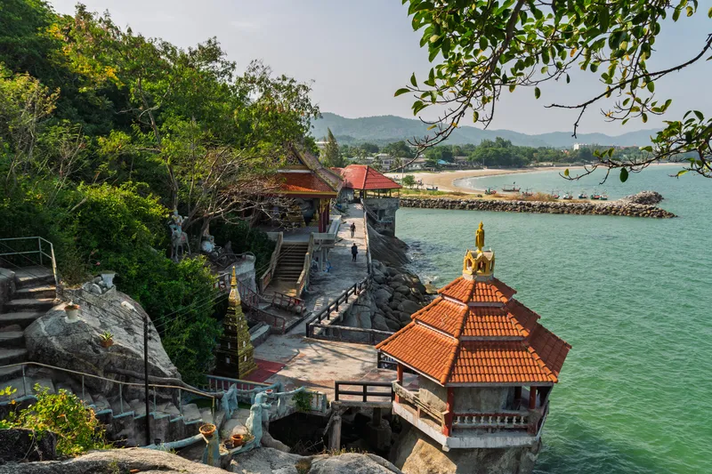 A seaside Buddhist temple complex built into rocky cliffs along a turquoise-green coastline, likely in Thailand. Orange-tiled roofed pavilions and shrines are perched on the rocks overlooking the calm sea. Stone staircases with metal railings wind up through the hillside, flanked by statues and small golden stupas. Lush tropical vegetation covers the hillside to the left, with mature trees providing canopy overhead. In the background, a sandy beach curves along the shore with fishing boats moored near a stone breakwater. Green mountains rise in the hazy distance under an overcast sky. A few visitors walk along the pathways between the temple structures. A golden Buddha statue stands atop one of the smaller shrine buildings near the water's edge.