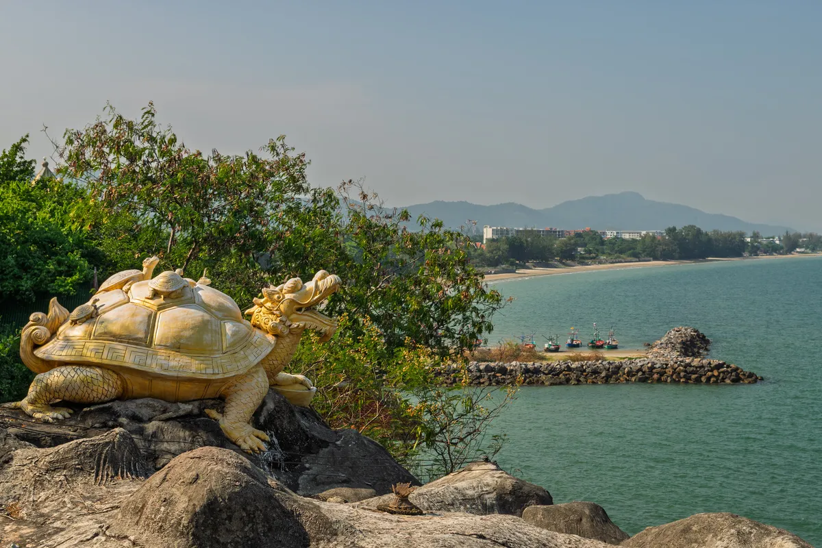 A large golden turtle sculpture with a dragon's head sits on a rocky outcrop by the sea. Several smaller turtles rest on the large turtle's shell. The ornately decorated statue bears traditional Asian patterns on the shell. In the background stretches a coastal landscape with turquoise water, a sandy beach, green trees, several brightly painted fishing boats at a stone jetty, and buildings and mountains outlined in the haze. The sky is slightly hazy and bright. The scene blends cultural symbolism with natural coastal beauty, likely in Thailand or Vietnam.