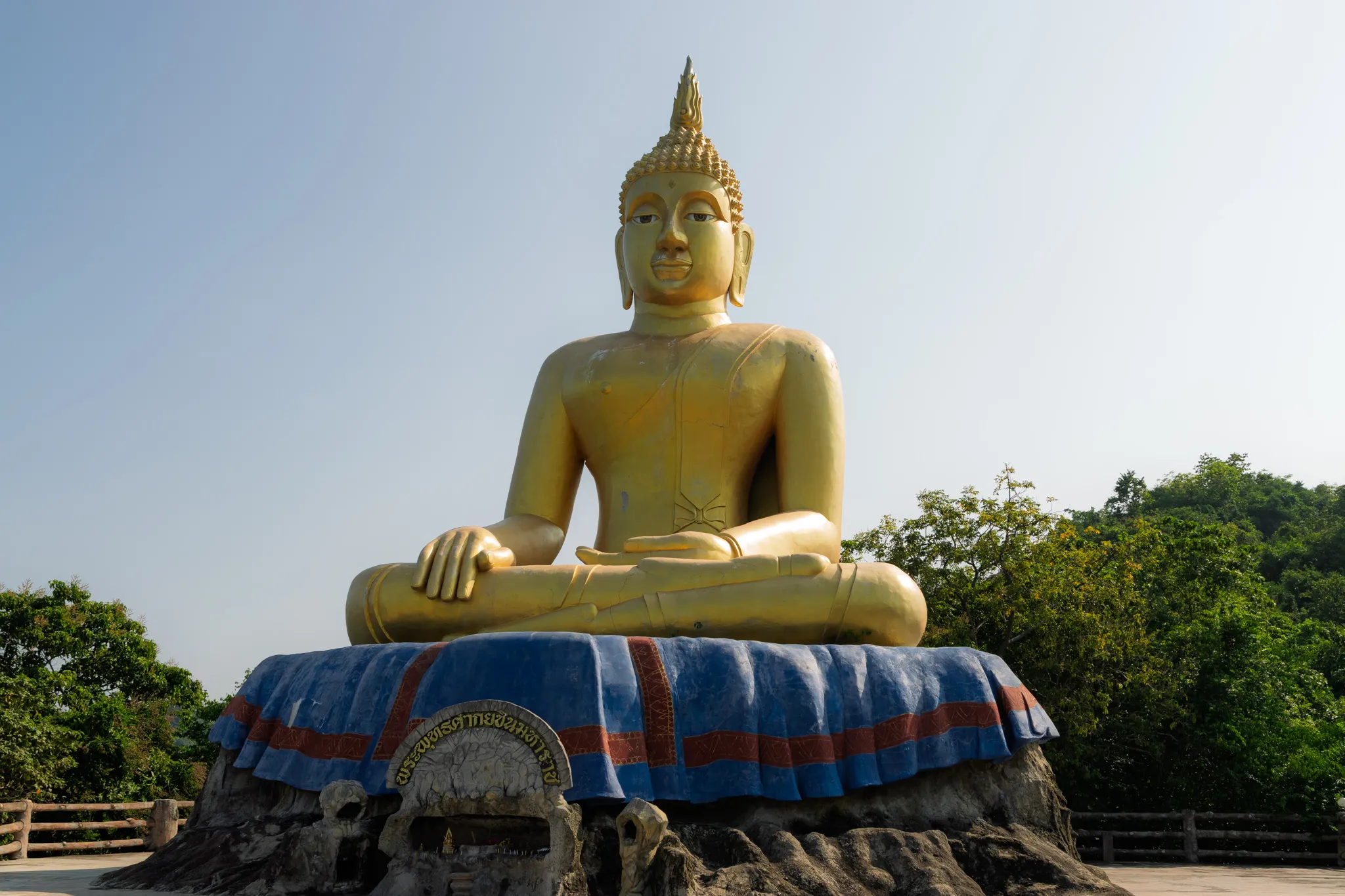 A large golden Buddha statue seated in the meditation posture (Bhumisparsha Mudra, with the right hand touching the earth) atop a rocky hilltop pedestal draped in blue and red fabric. The statue features a pointed flame-like ushnisha crown, elongated earlobes, and a serene facial expression characteristic of Thai Buddhist art. At the base of the pedestal, there is a circular plaque with Thai script. The statue is surrounded by lush green tropical trees, with a hazy pale blue sky in the background. A railing is visible on the left side, suggesting this is a viewpoint or elevated temple site, likely in Thailand.