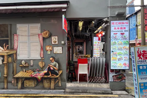 In Tanjong Pagar, Outram, Central Region, Singapore, a small alley features a richly detailed mural depicting a traditional craft. On the left, an elderly man sits on a wooden board, sewing a colourful piece of fabric. Beside him are various objects such as baskets and old pictures hanging on a wall. The entrance leads to a restaurant with red and white chairs on a paved floor. Multilingual menus on the wall list a variety of dishes. The street is marked with yellow lines creating parking space.