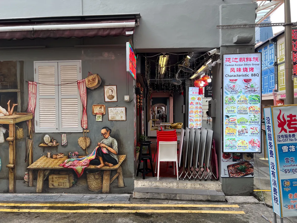 In Tanjong Pagar, Outram, Central Region, Singapore, a small alley features a richly detailed mural depicting a traditional craft. On the left, an elderly man sits on a wooden board, sewing a colourful piece of fabric. Beside him are various objects such as baskets and old pictures hanging on a wall. The entrance leads to a restaurant with red and white chairs on a paved floor. Multilingual menus on the wall list a variety of dishes. The street is marked with yellow lines creating parking space.