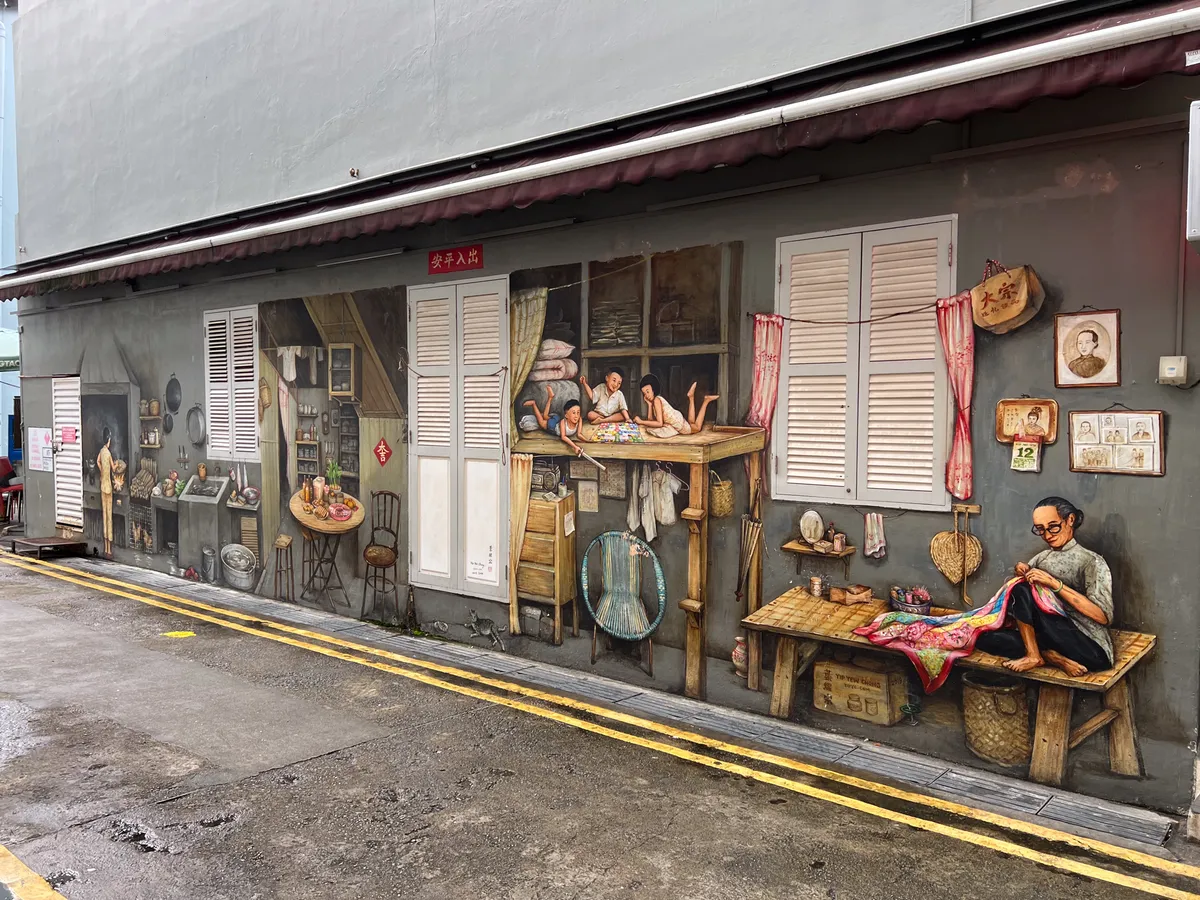 In Tanjong Pagar, Outram, Central Region, Singapore, a lively mural on a wall depicts a traditional craft and everyday scene. On the left, a man appears in a kitchen scene surrounded by pots and food. At a table, two women sit sewing together. On the right, an elderly woman is shown concentrating on a colourful fabric. Behind her, shelves with various utensils and pictures on the wall create a nostalgic atmosphere. The scene captures details of the old way of life in Singapore.