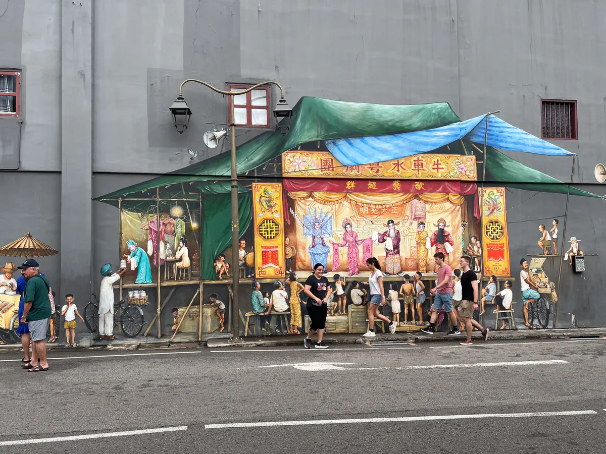 In Tanjong Pagar, Outram, Central Region, Singapore, a colourful mural depicts a lively market scene. On the right, brightly dressed figures stand in a festive square, while in the foreground people of all ages are engaged in various activities such as eating, drinking and shopping. The scene feels busy and dynamic, with a tent covering the space and countless details reflecting everyday life and the culture of the region.
