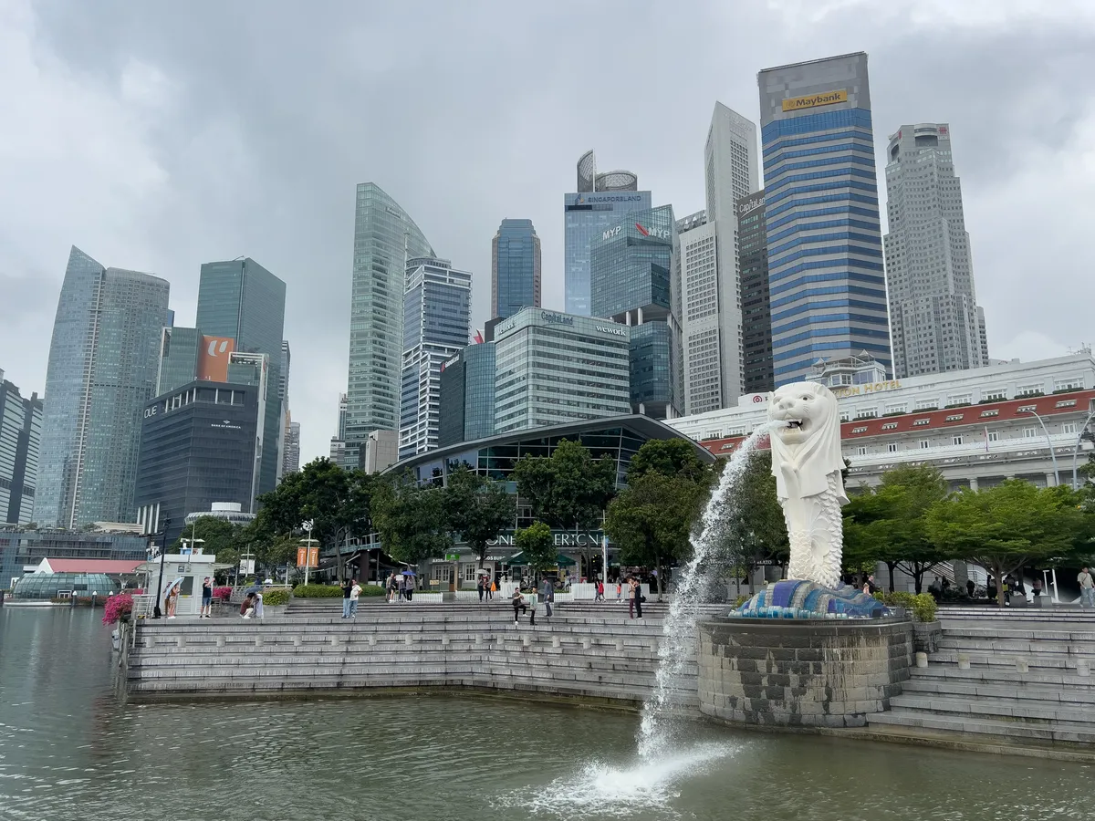 In the Civic District, Downtown Core, Central Region, Singapore, 179803, the Merlion stands in all its splendour, an iconic statue spouting water from its mouth. Surrounded by modern skyscrapers under a cloudy sky, the scene blends nature with urban architecture. In the background, high-rises with reflective windows and green trees bring the square to life. Visitors stroll along the promenade while the Merlion draws all the attention.