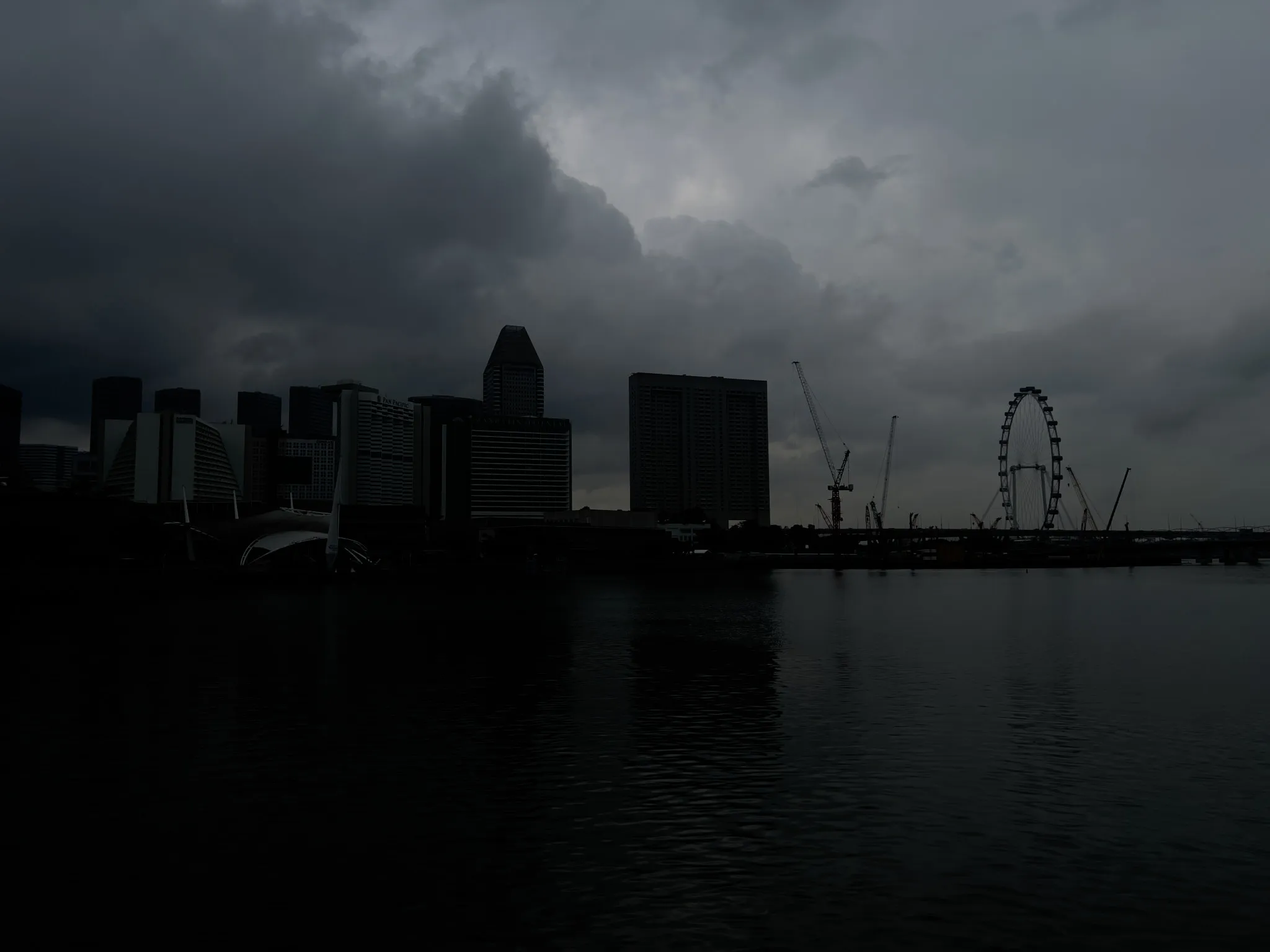 A dark, moody waterfront scene showing a city skyline silhouetted beneath heavy storm clouds, with tall modern buildings, construction cranes, and a large Ferris wheel visible across the water. The surface of the water reflects the dim light from the overcast sky, adding to the subdued, dramatic atmosphere.