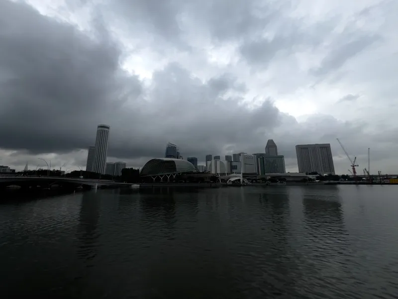 A waterfront city skyline under heavy, overcast clouds with dark, dramatic shading. A calm body of water fills the foreground, reflecting the muted shapes of tall buildings, a domed structure, cranes, and a bridge stretching across the left side. The atmosphere appears moody and dim, suggesting impending rain as thick cloud cover dominates the sky.