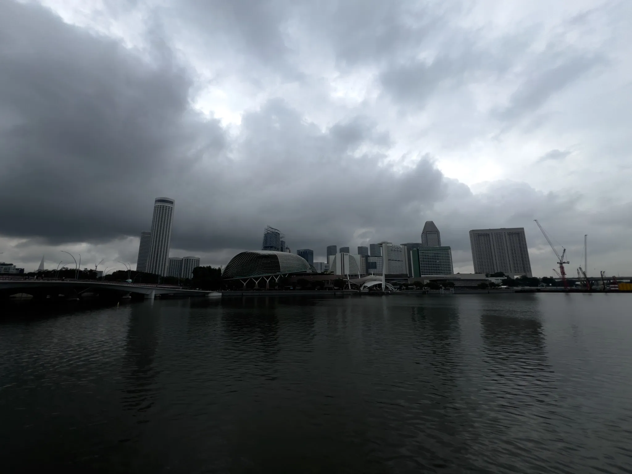 A waterfront city skyline under heavy, overcast clouds with dark, dramatic shading. A calm body of water fills the foreground, reflecting the muted shapes of tall buildings, a domed structure, cranes, and a bridge stretching across the left side. The atmosphere appears moody and dim, suggesting impending rain as thick cloud cover dominates the sky.