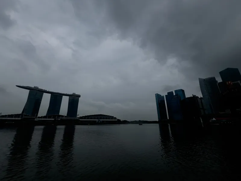 A dramatic urban waterfront scene under a heavy, overcast sky with dense, dark clouds. Distinctive modern skyscrapers rise on the right side, while three tall towers supporting a long, curved structure stand prominently on the left. Their reflections ripple faintly across the calm water in the foreground, creating a moody and atmospheric view of the cityscape.