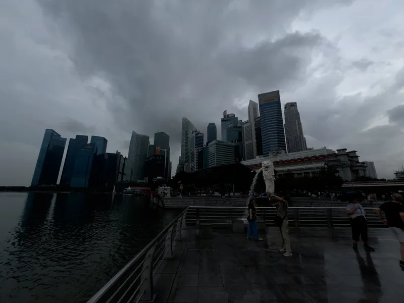 A waterfront promenade facing a dense cluster of tall modern skyscrapers under a dark, overcast sky. The Merlion statue stands prominently near the center, spraying water into the bay. The wet pavement reflects the cloudy atmosphere, and several people are scattered around the viewing area, some taking photos. The water is calm, mirroring the outline of the buildings on the left, while the overall scene feels moody and dramatic due to the heavy clouds.