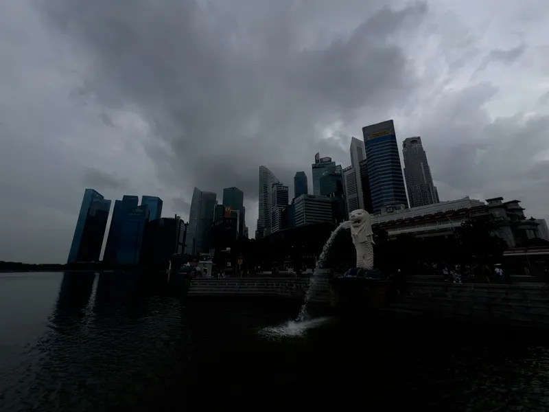 The Merlion statue stands at the edge of the waterfront, spouting water into the bay beneath a dark, overcast sky. Tall modern skyscrapers rise closely behind it, their glass facades dim under the heavy, gray clouds. The water reflects the muted tones of the scene, while small groups of people gather near the promenade in the distance.