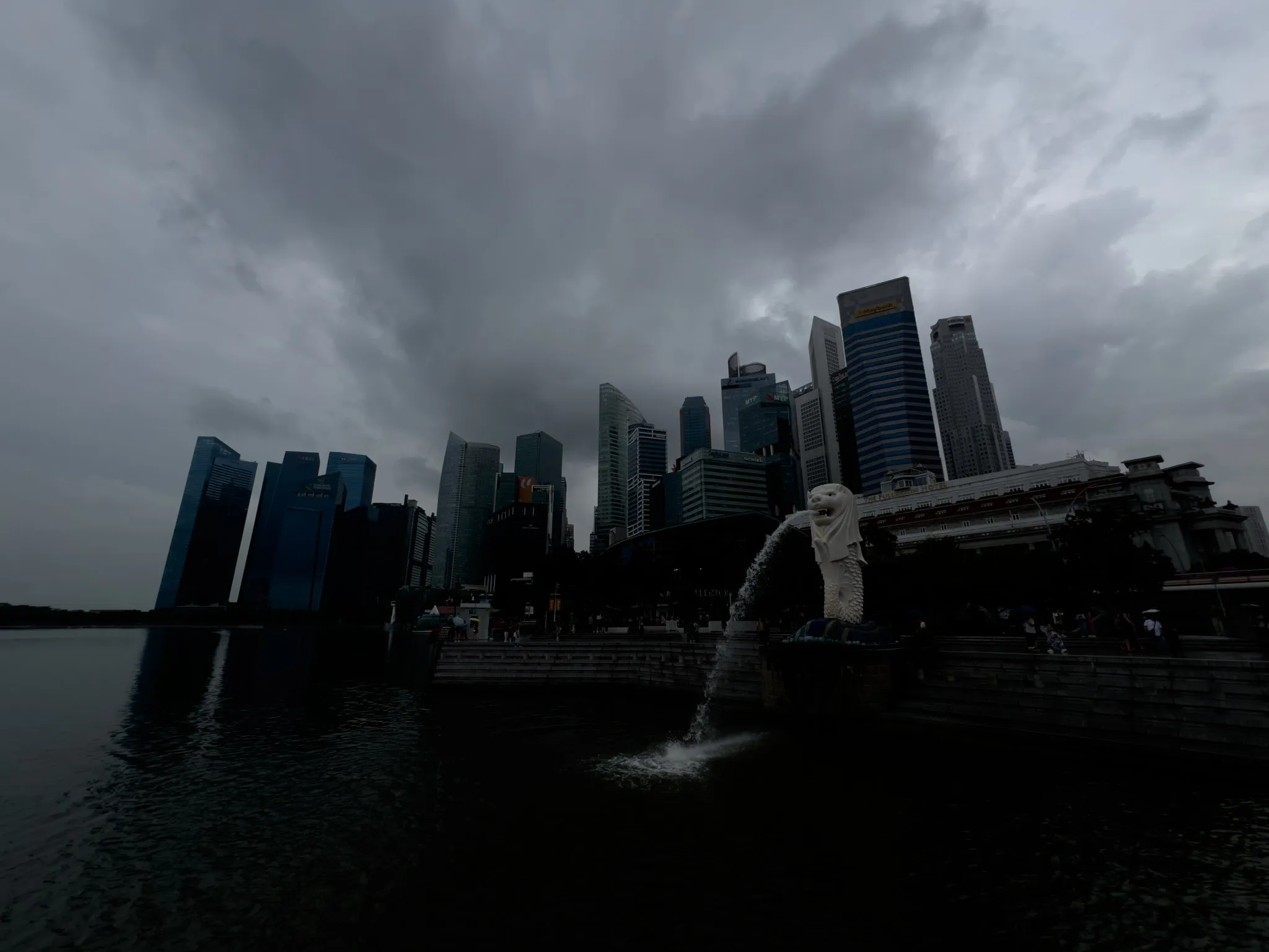 The Merlion statue stands at the edge of the waterfront, spouting water into the bay beneath a dark, overcast sky. Tall modern skyscrapers rise closely behind it, their glass facades dim under the heavy, gray clouds. The water reflects the muted tones of the scene, while small groups of people gather near the promenade in the distance.