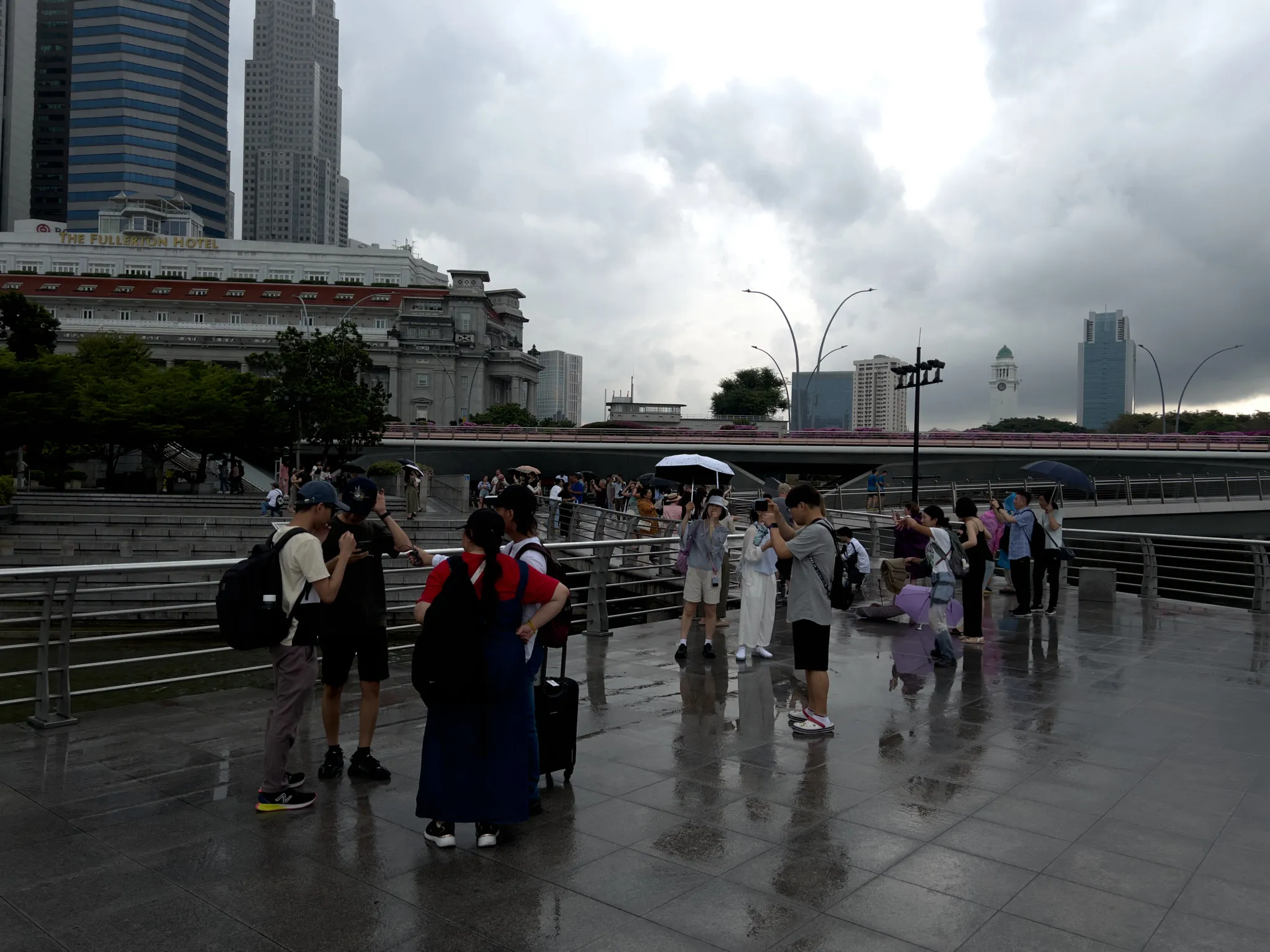 A busy waterfront promenade with groups of people standing and walking on a wet, reflective stone surface after rainfall. Several individuals hold umbrellas while others take photos or chat in small groups. Modern high-rise buildings and an older, ornate structure with red awnings form the backdrop. A bridge stretches across the mid-ground, with more people visible along its walkway. The sky is filled with heavy gray clouds, suggesting lingering or impending rain.