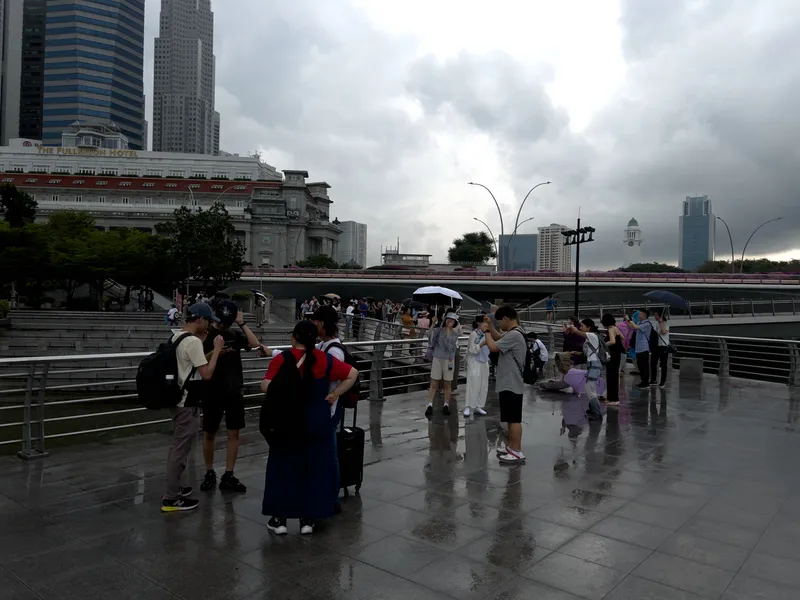 A busy waterfront promenade with groups of people standing and walking on a wet, reflective stone surface after rainfall. Several individuals hold umbrellas while others take photos or chat in small groups. Modern high-rise buildings and an older, ornate structure with red awnings form the backdrop. A bridge stretches across the mid-ground, with more people visible along its walkway. The sky is filled with heavy gray clouds, suggesting lingering or impending rain.