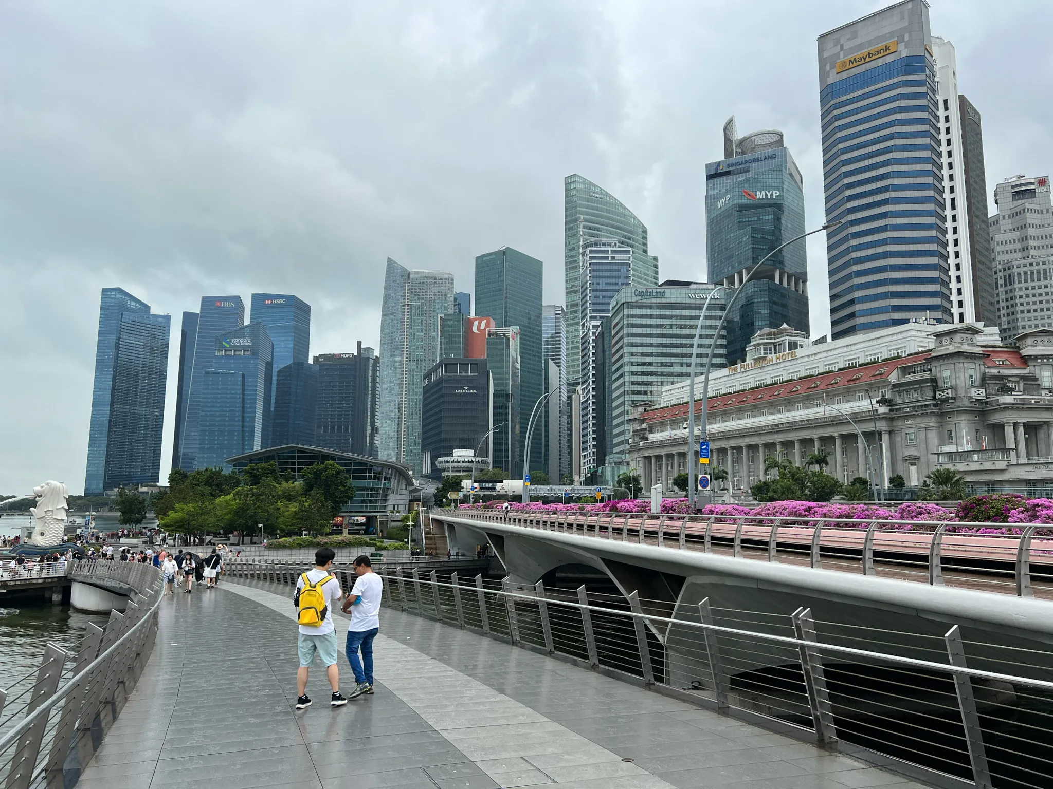 A waterfront promenade curves alongside the bay, where people walk near steel railings and wet pavement under an overcast sky. A white Merlion statue stands in the distance on the left, facing the water. Across the bay rises a dense cluster of tall modern skyscrapers with glass facades, while a large historic-style building with a red roof sits prominently on the right next to a bridge lined with bright pink flowers. The overall scene blends contemporary architecture, greenery, and pedestrian activity in a busy urban setting.