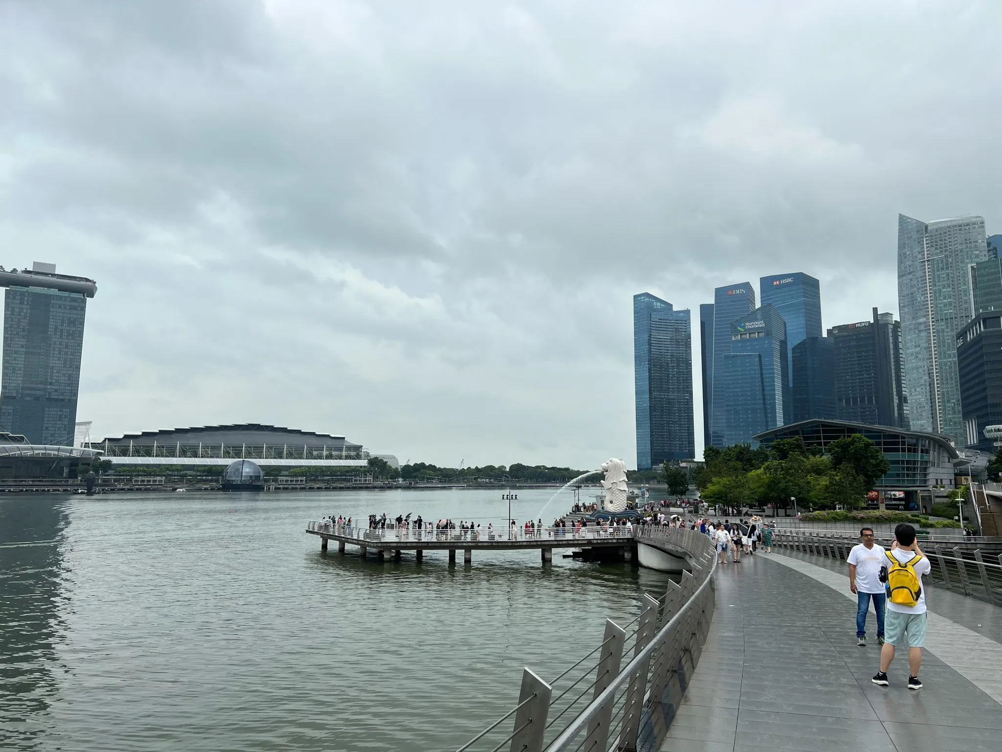 Wide waterfront promenade curving along the bay with people walking and taking photos, leading toward a platform where a white fountain statue sprays water into the bay. Tall modern skyscrapers rise on the right side behind trees, while distinctive architectural structures sit across the water on the left. The sky is filled with thick gray clouds, casting diffused light over the calm water and the bustling walkway.